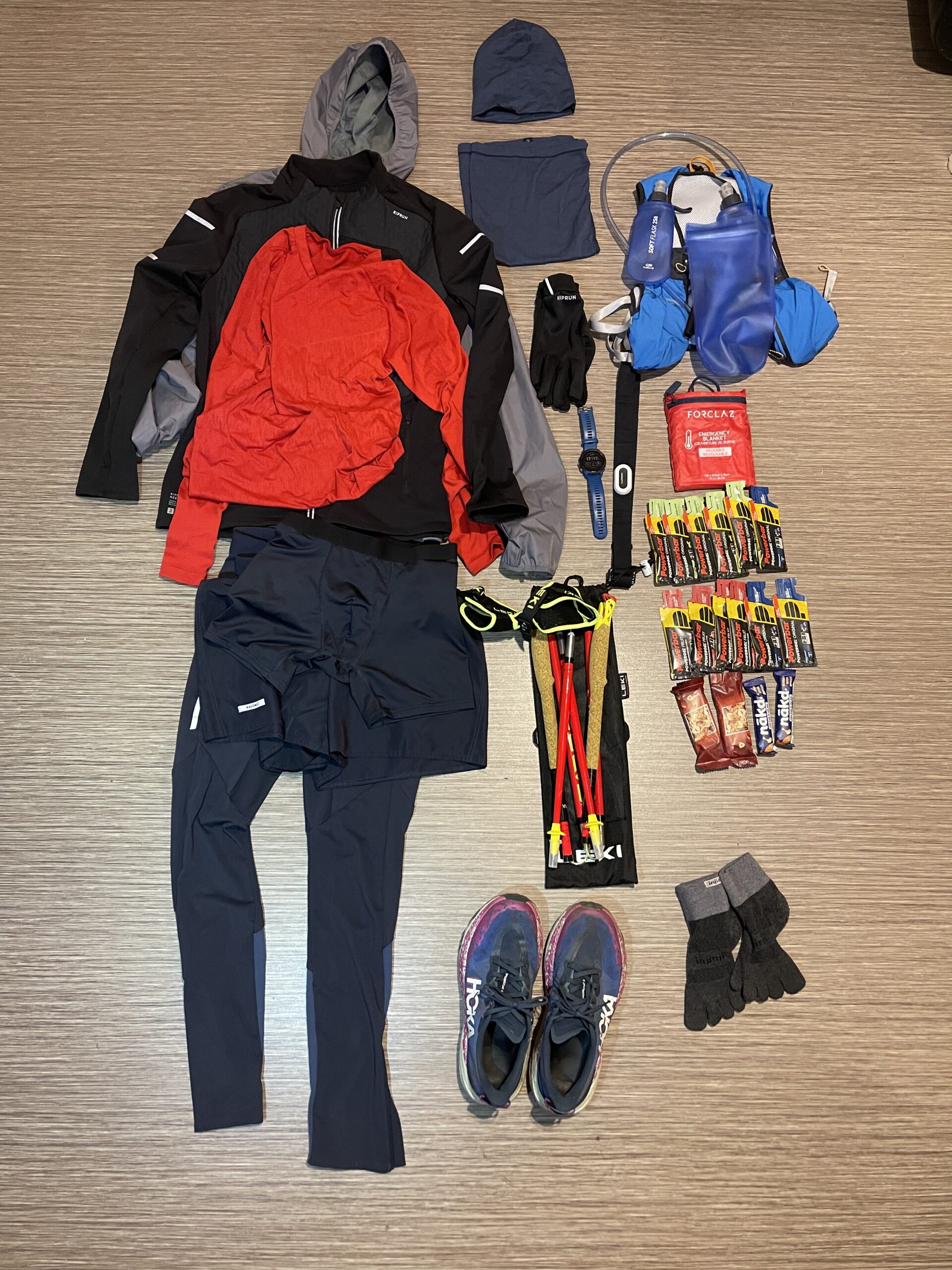 Flat lay of winter trail running gear before a 27 km race, including trail shoes, gloves, hydration vest, soft flasks, poles, watch, heart rate monitor, emergency blanket, and gels.