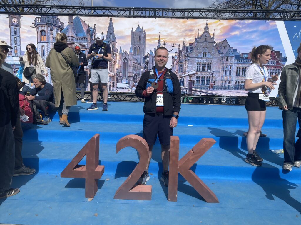 Proud runner standing behind large 42 numbers after completing first marathon training race, with race banners and other finishers visible in background.