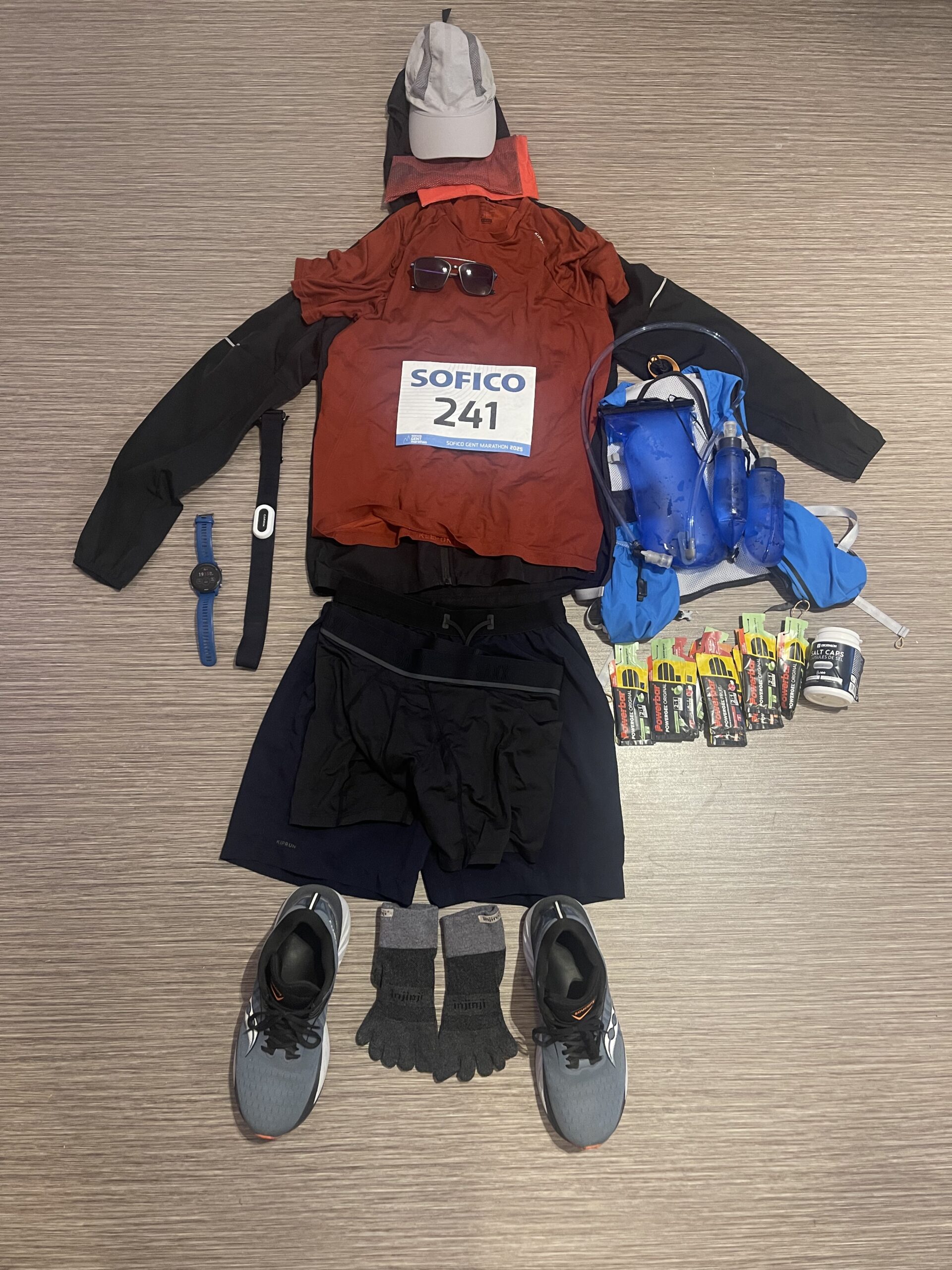 Flat lay of complete marathon running gear including clothing, hydration vest, nutrition, GPS watch, heart rate monitor, shoes, and race bib for first marathon test race.