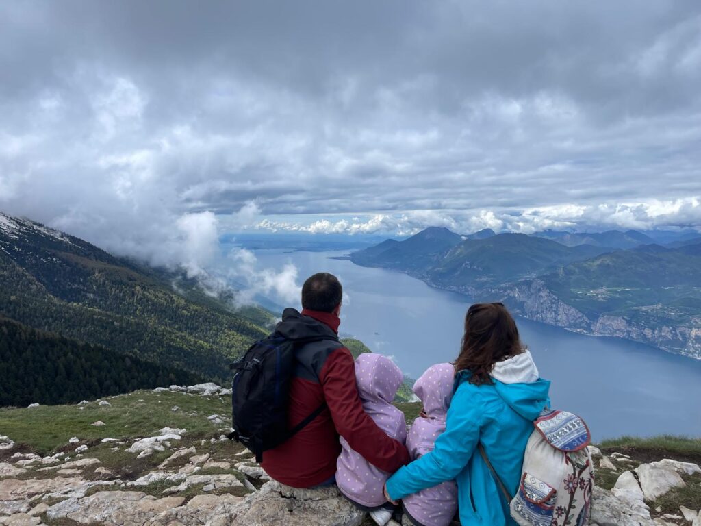 Family sitting on a mountaintop above Lago di Garda, hugging with a panoramic view of the lake and surrounding mountains.