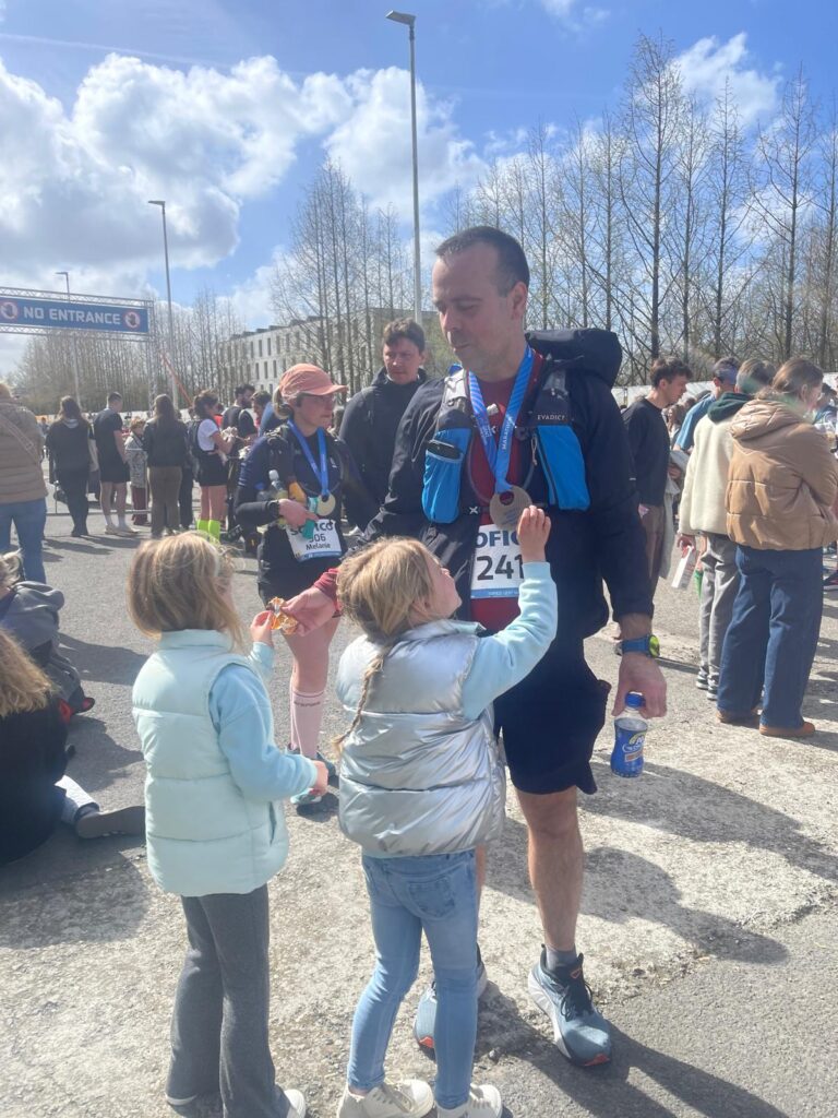 Loránd after his first marathon, wearing a medal, sharing finish line snacks with one daughter while the other admires the medal.