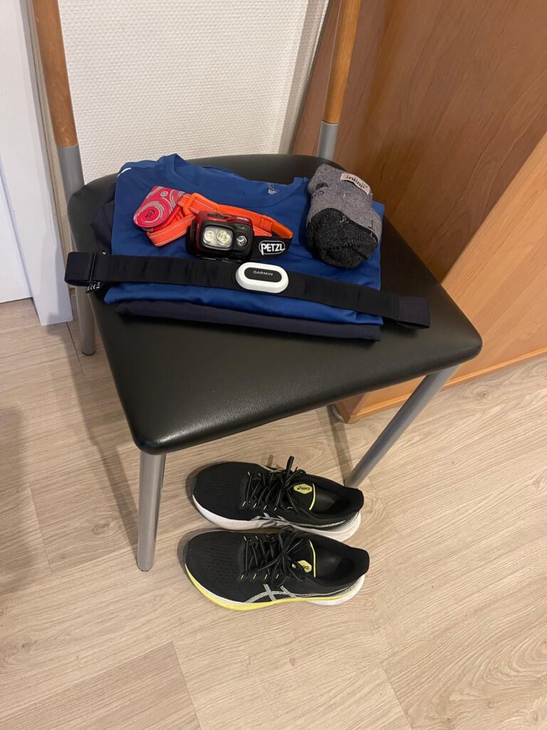 Folded running gear on a chair, heart rate monitor and headlamp ready, and running shoes placed under the chair, prepared for the next morning’s run