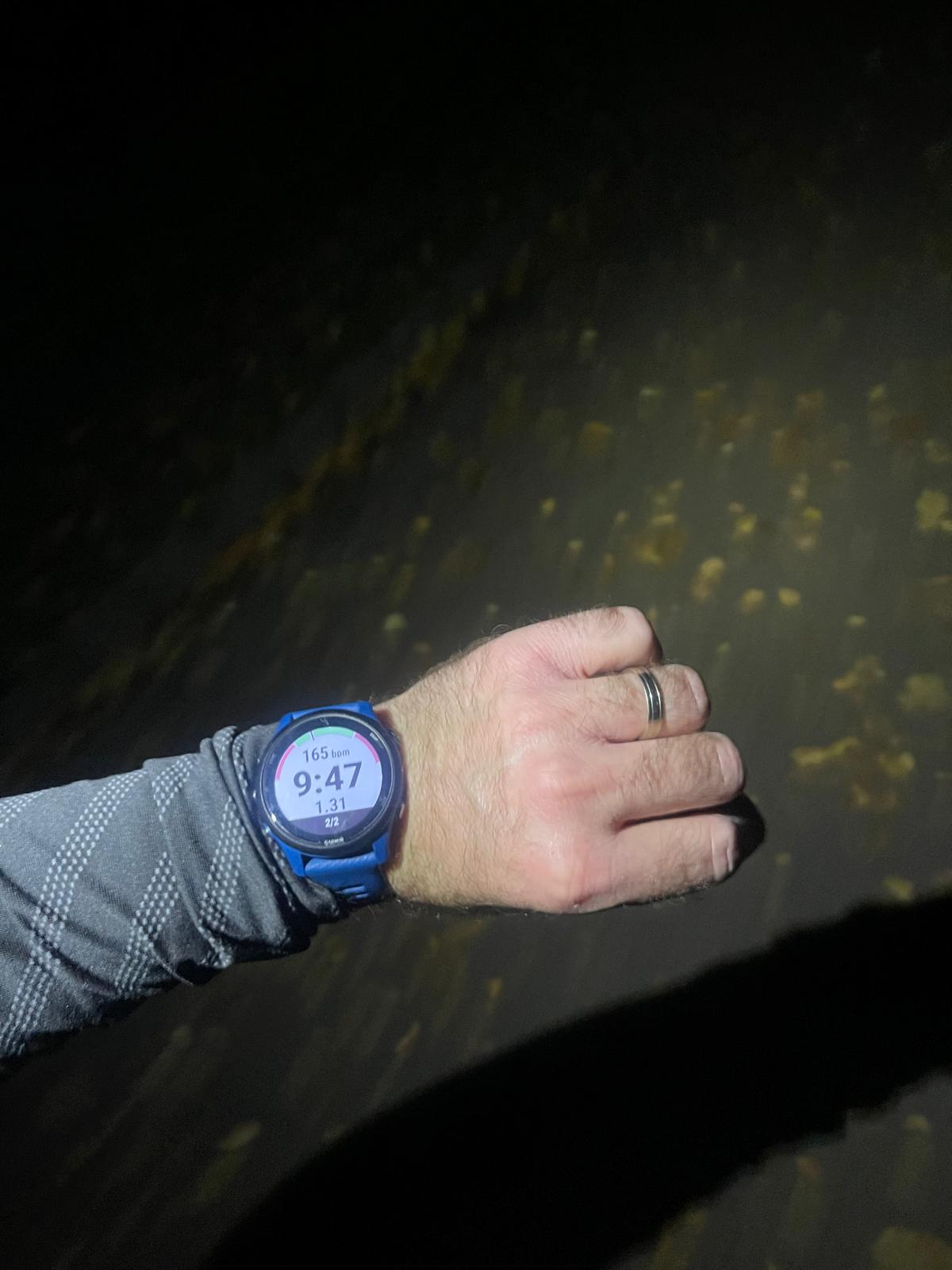 Photo taken mid-run in the dark, showing the illuminated screen of a Garmin Forerunner 255 watch displaying threshold workout metrics. A headtorch lights up the asphalt path in the park, revealing autumn leaves and the runner’s arm.