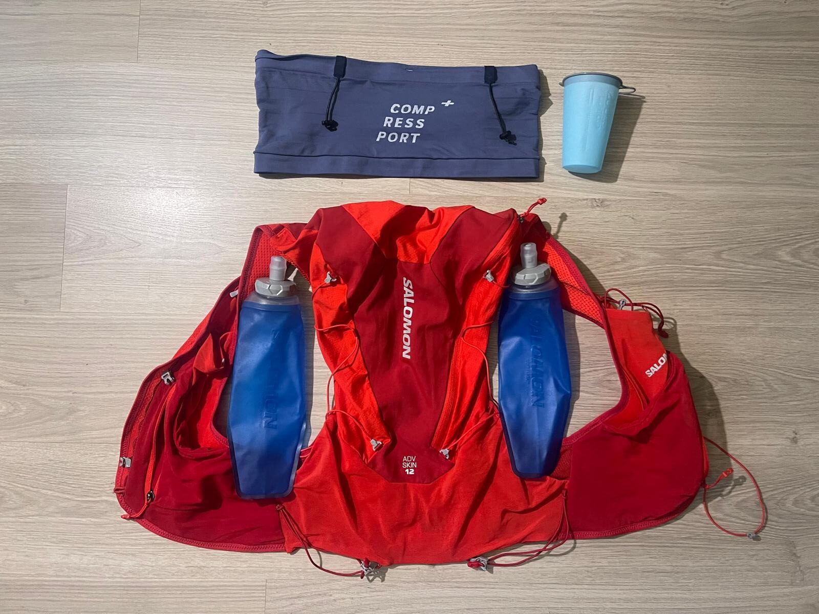 Flat lay photo of hydration setup with Compressport running belt, Salomon hydration vest, soft flasks, and foldable running cup.