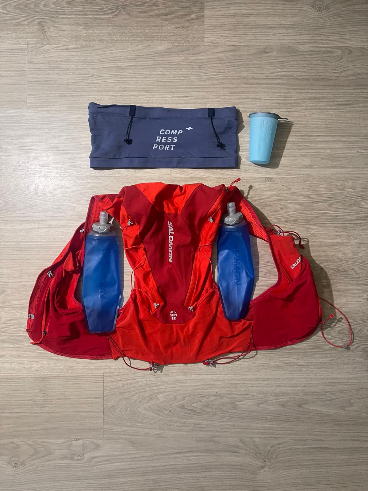 Flat lay photo of hydration setup with Compressport running belt, Salomon hydration vest, soft flasks, and foldable running cup.