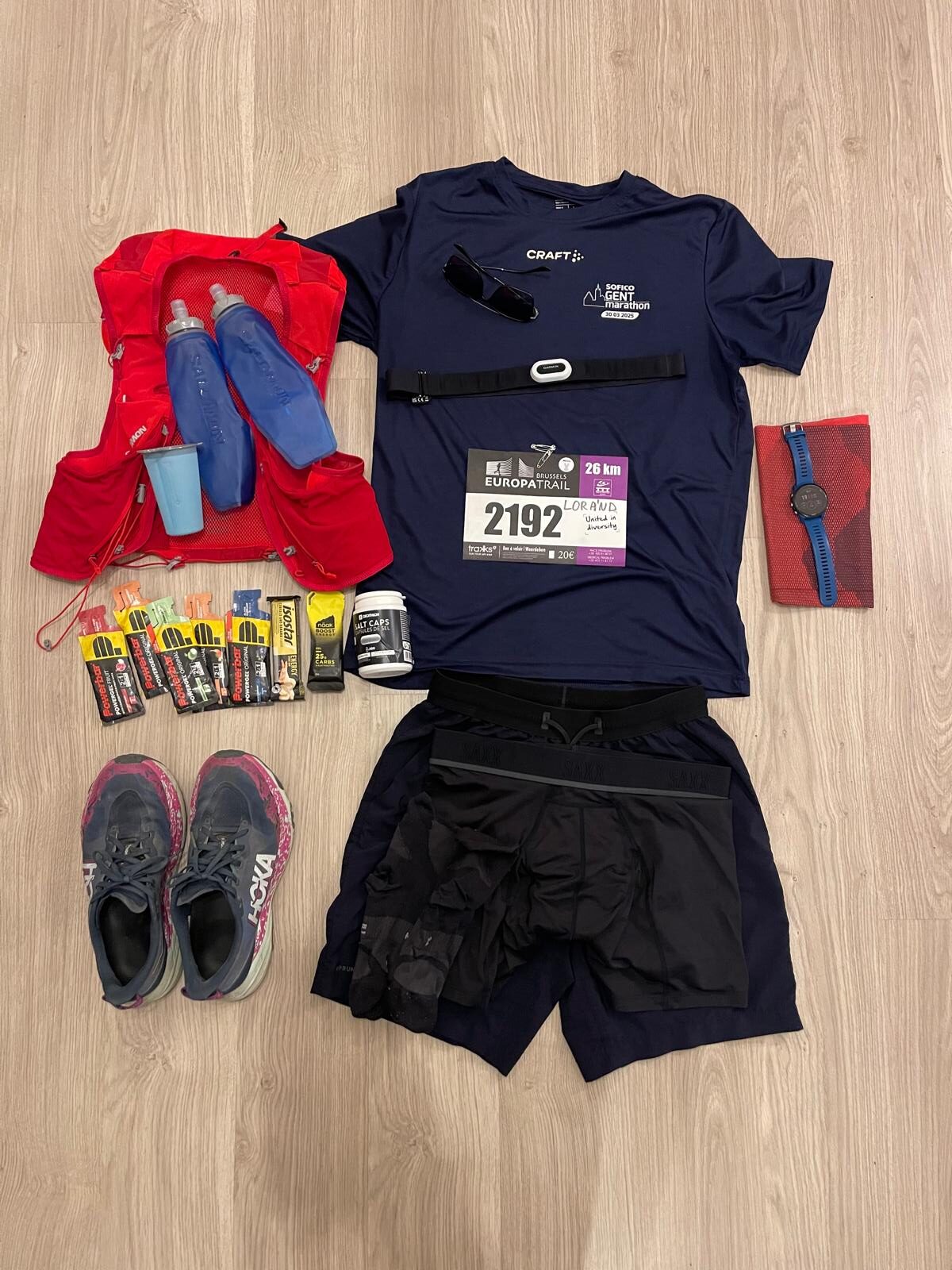 Flat lay of gear and clothing for first UTMB index trail run including Salomon Adv Skin 12 hydration vest, soft flasks, foldable cup, Hoka Speedgoat 6, energy gel, salt tablets, Garmin HRM Pro Plus, Garmin Forerunner 255, shorts, shirt, glasses, buff, sport boxers, socks and race bib number placed on the shirt