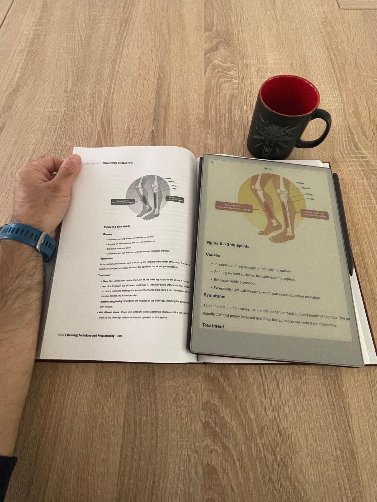 Study session for ISSA Running Coach certification — open course material on the table, Remarkable Paper Pro tablet showing a zoomed-in illustration from the same book, Witcher coffee mug on the right, Garmin running watch visible on left wrist.